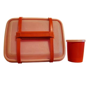Puede incluir: Un juego de fiambrera Tupperware vintage en un tono rosa claro, asegurado con correas de color naranja brillante y una taza naranja a juego. La fiambrera tiene el logotipo de Tupperware en relieve en el asa naranja.