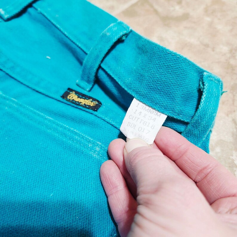 May include: Close-up of turquoise Wrangler jeans. The jeans have a classic five-pocket design and a black and gold Wrangler label. A white tag with text is held by a hand. The jeans are a vibrant shade of turquoise.