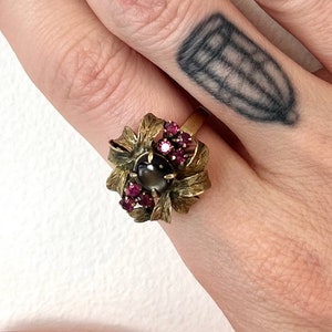 May include: A gold ring with a dark gray stone surrounded by red stones set in a floral design.