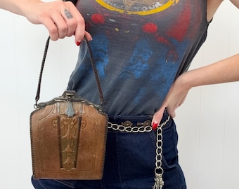 May include: A brown leather handbag with a decorative metal clasp and handle. The bag has a rectangular shape with embossed detailing. The person is wearing a gray graphic t-shirt and denim shorts.