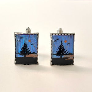 May include: A pair of cufflinks featuring a blue and orange butterfly wing design with a silhouette of a tree and a setting sun. The cufflinks are silver-toned and have a decorative border.
