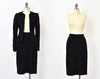 May include: A black skirt suit with a fitted jacket and a straight skirt. The jacket has a collar and long sleeves. The skirt is knee-length.