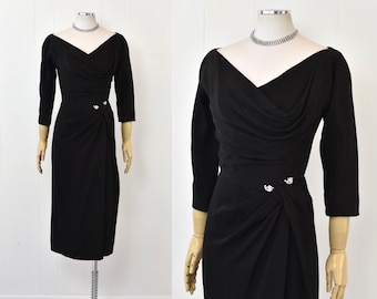 May include: A black, knee-length dress with a draped neckline and three-quarter sleeves. The dress has a fitted silhouette and features a decorative button detail at the waist.