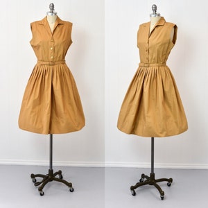 May include: A sleeveless, collared dress with a button-up front and a belt. The dress is a light brown color and has a pleated skirt.
