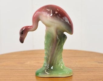 May include: A pink and white ceramic flamingo figurine standing on a green base. The flamingo is posed with its head bent down, as if it is drinking water.