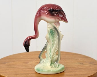 May include: A pink ceramic flamingo figurine standing on a green and white base. The flamingo is in a standing position with its head bent down.
