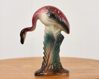 May include: A pink and white ceramic flamingo figurine standing on a green and brown base. The flamingo is in a bent-over pose, with its head facing down.