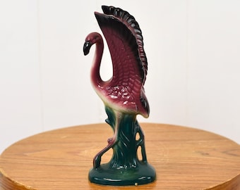 May include: A ceramic flamingo figurine with a pink and purple body and black wings. The flamingo is standing on a green base.
