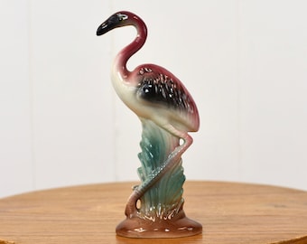 May include: A ceramic flamingo figurine with pink, black, and green paint. The flamingo is standing on a green base with brown accents.