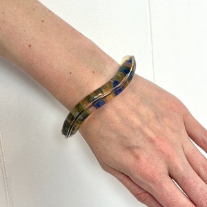 May include: A wavy, multi-colored glass bangle bracelet with blue, yellow, and brown tones. The bracelet is open and has a smooth, polished finish.