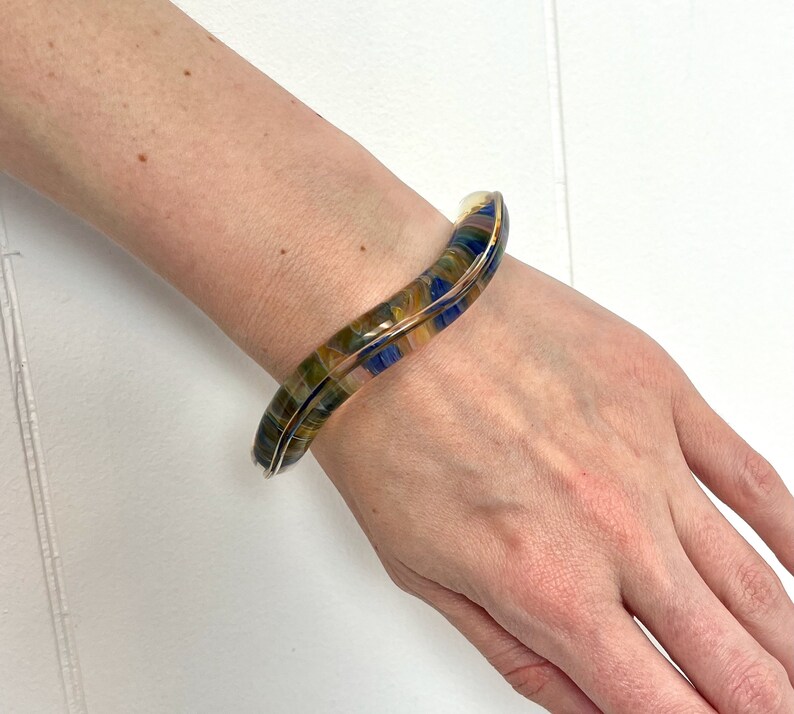 May include: A wavy, multi-colored glass bangle bracelet with blue, yellow, and brown tones. The bracelet is open and has a smooth, polished finish.