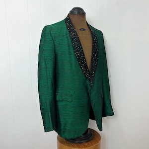 Może przedstawiać: Zielony blazer z czarnym i złotym kołnierzem z cekin&oacute;w. Blazer wykonany jest z fakturowanej tkaniny i ma pojedyncze zapięcie na guzik.