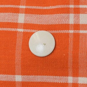 May include: A small, off-white, round button with two holes is centered on an orange and white plaid fabric. The button is smooth and appears to be made of a solid material. The fabric has a woven texture.