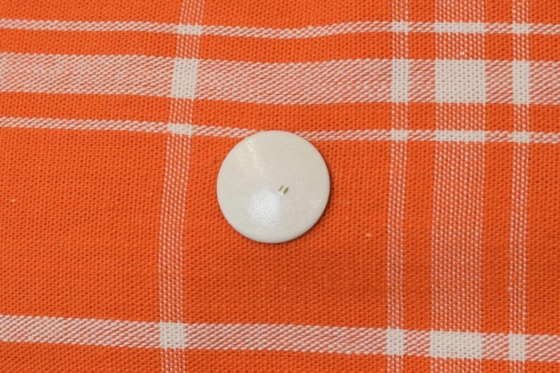 May include: A small, off-white, round button with two holes is centered on an orange and white plaid fabric. The button is smooth and appears to be made of a solid material. The fabric has a woven texture.