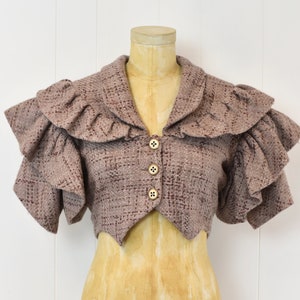 May include: A cropped brown tweed jacket with a ruffled collar and sleeves. The jacket has a pointed hem and is fastened with four buttons.