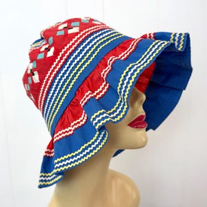 May include: A red and blue sun hat featuring a geometric pattern on the crown with small white and light blue squares. The wide brim is accented with multiple rows of blue fabric ruffles, each trimmed with a wavy white and yellow ribbon edge.