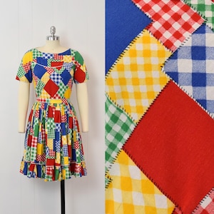 May include: A colorful patchwork dress with short sleeves and a flared skirt. The dress features a variety of patterns, including gingham and diamond shapes, in red, yellow, green, and blue. The dress is displayed on a mannequin.