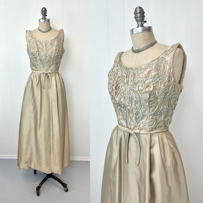 Op de afbeelding: Een vintage, mouwloze jurk met een getailleerd lijfje en een volle rok. Het lijfje is versierd met zilveren pailletten en kralen in een bloemenpatroon. De jurk is licht goud van kleur.