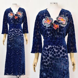 May include: A floor-length navy blue velvet dress featuring a burnout floral pattern, three-quarter length sleeves, a V-neckline, and a wide waistband. The bodice is adorned with three large, dimensional fabric flowers in shades of blue and pink.