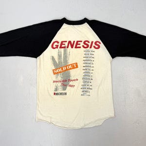 May include: Cream-colored raglan shirt with black sleeves. The shirt features the band name "Genesis" in red, a hand graphic, and tour details for the "Invisible Touch Tour 1987". The shirt also has the text "SOLD OUT".