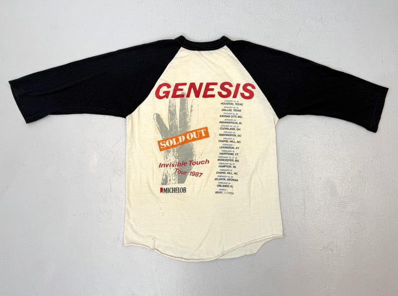 May include: Cream-colored raglan shirt with black sleeves. The shirt features the band name "Genesis" in red, a hand graphic, and tour details for the "Invisible Touch Tour 1987". The shirt also has the text "SOLD OUT".