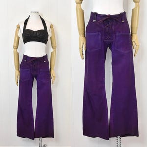May include: A pair of vintage purple suede pants with a flared leg. The pants feature a lace-up front detail and two back pockets. The pants are displayed on a mannequin with a black halter top.