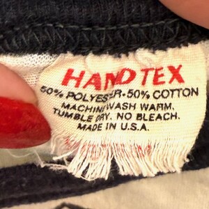 May include: Close-up of a fabric tag with the text "HAND TEX" in red. The tag also includes washing instructions and the text "MADE IN U.S.A." The tag is made of a cream-colored fabric.
