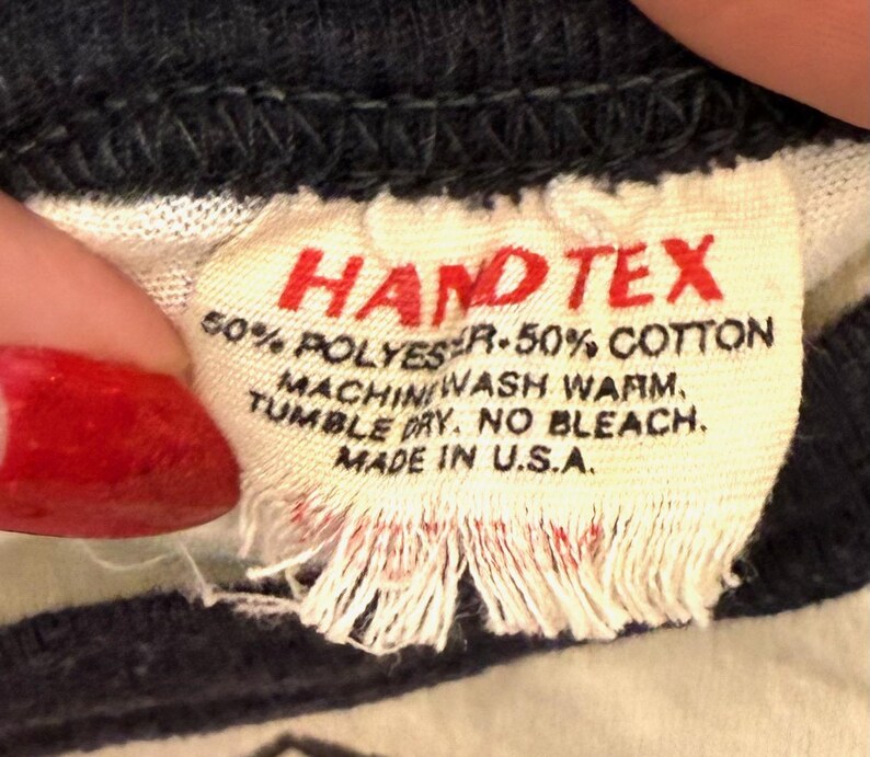 May include: Close-up of a fabric tag with the text "HAND TEX" in red. The tag also includes washing instructions and the text "MADE IN U.S.A." The tag is made of a cream-colored fabric.