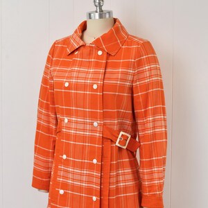 May include: An orange plaid coat with white accents, displayed on a mannequin. The coat features a double-breasted design with white buttons, a matching belt with a gold buckle, and a classic collar. The long sleeves and tailored fit suggest a vintage style.