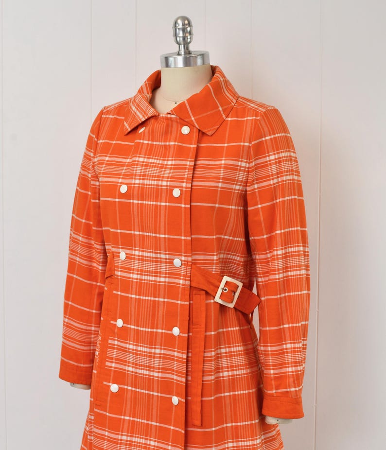 May include: An orange plaid coat with white accents, displayed on a mannequin. The coat features a double-breasted design with white buttons, a matching belt with a gold buckle, and a classic collar. The long sleeves and tailored fit suggest a vintage style.