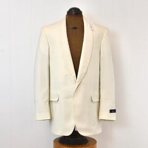 May include: Cream-colored tuxedo jacket with a shawl collar, two front pockets, and a single-button closure. The jacket is displayed on a mannequin. A small tag is attached to the lapel. The jacket is made of a smooth fabric.