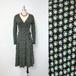 May include: A black wrap dress with a floral pattern in light blue and green. The long-sleeved dress has a V-neck and a tie at the waist. The dress is displayed on a mannequin.