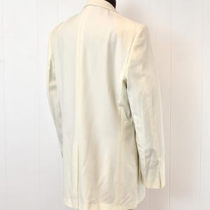 May include: A light cream-colored blazer displayed on a mannequin. The blazer has a classic design with notched lapels, a two-button closure, and a tailored fit. The sleeves have buttoned cuffs. The garment is made of a smooth fabric and is suitable for formal or semi-formal occasions.