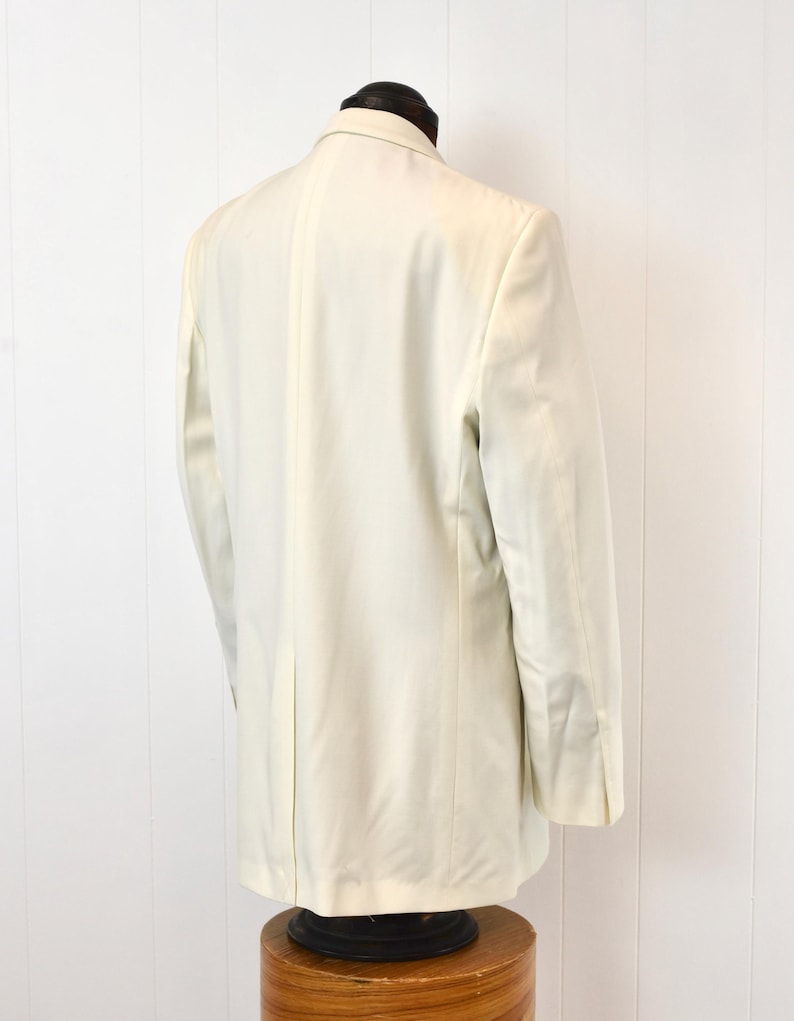 May include: A light cream-colored blazer displayed on a mannequin. The blazer has a classic design with notched lapels, a two-button closure, and a tailored fit. The sleeves have buttoned cuffs. The garment is made of a smooth fabric and is suitable for formal or semi-formal occasions.