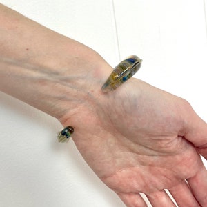 May include: A clear glass bangle bracelet with swirls of blue, green, and yellow. The bracelet is open and has a slightly curved shape.