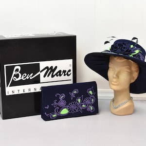 May include: A navy blue hat with a green and purple floral design and a matching navy blue clutch with a green and purple floral design. The hat is on a mannequin head and the clutch is on a black box with the text "Ben Marc International" on it.