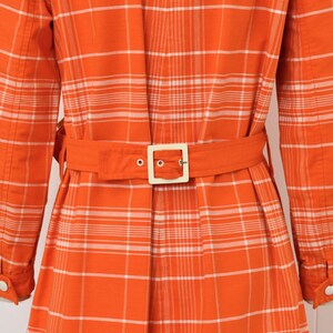 May include: Orange plaid coat with a matching belt and a square buckle. The long-sleeved coat has white buttons and a classic collar. The plaid pattern is orange and white.