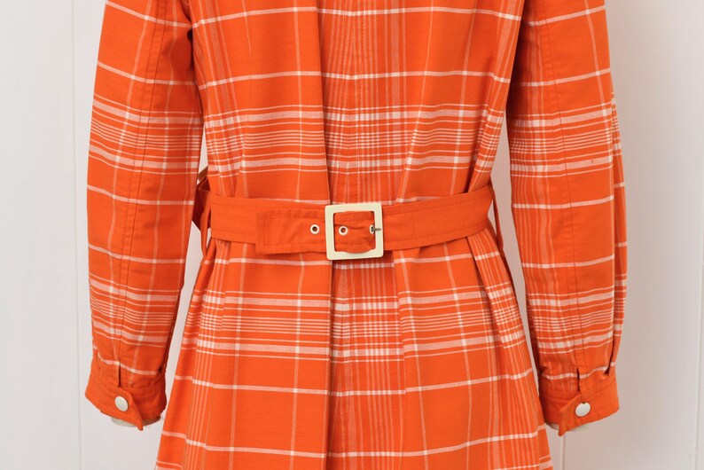 May include: Orange plaid coat with a matching belt and a square buckle. The long-sleeved coat has white buttons and a classic collar. The plaid pattern is orange and white.