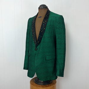 Może przedstawiać: Zielony blazer z czarnym aksamitnym kołnierzem ozdobionym złotymi i zielonymi cekinami. Blazer ma pojedyncze zapięcie na guzik i kieszeń po lewej stronie.