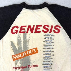 May include: Cream-colored vintage t-shirt with black sleeves, featuring the band name "GENESIS" in red block letters. The shirt has a hand graphic and a "SOLD OUT" sticker. Tour dates are listed below.