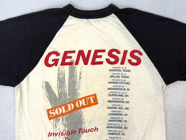 May include: Cream-colored vintage t-shirt with black sleeves, featuring the band name "GENESIS" in red block letters. The shirt has a hand graphic and a "SOLD OUT" sticker. Tour dates are listed below.