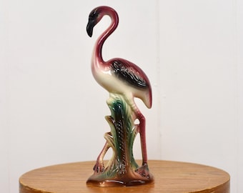 May include: A ceramic flamingo figurine with a curved neck and a black beak. The flamingo has pink, white, and black feathers, and stands on a green and brown base. The figurine is displayed on a wooden surface.