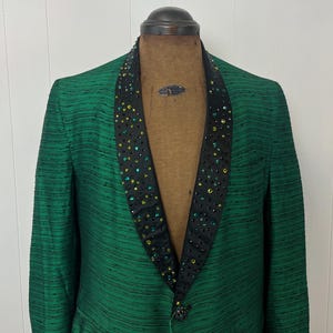 Może przedstawiać: Zielony blazer z czarnym satynowym kołnierzem ozdobionym zielonymi i niebieskimi kryształami. Blazer ma prążkowany wz&oacute;r i zapięcie na jeden guzik.