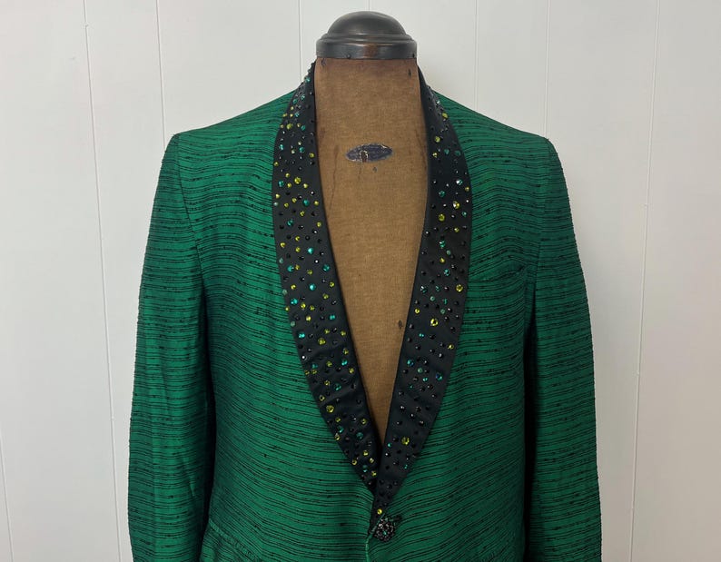 Może przedstawiać: Zielony blazer z czarnym satynowym kołnierzem ozdobionym zielonymi i niebieskimi kryształami. Blazer ma prążkowany wz&oacute;r i zapięcie na jeden guzik.