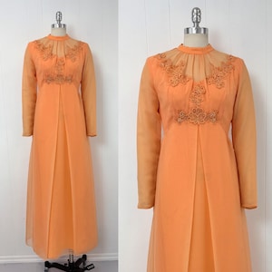 May include: A vintage orange maxi dress on a mannequin. The dress features long sheer sleeves, a high neckline, and intricate gold-colored embroidery on the bodice. The skirt is a flowing, solid orange fabric.