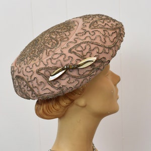 May include: A pink felt hat with a delicate gold and silver beaded design. The hat has a small, pearl-like brooch with a gold leaf design.