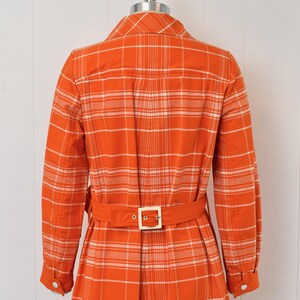 May include: An orange plaid dress with long sleeves and a matching belt. The dress has a collared neckline and a button-down front. The belt has a square, gold-colored buckle. The dress is displayed on a mannequin.