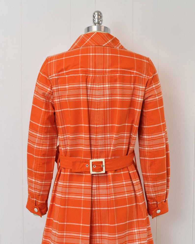 May include: An orange plaid dress with long sleeves and a matching belt. The dress has a collared neckline and a button-down front. The belt has a square, gold-colored buckle. The dress is displayed on a mannequin.