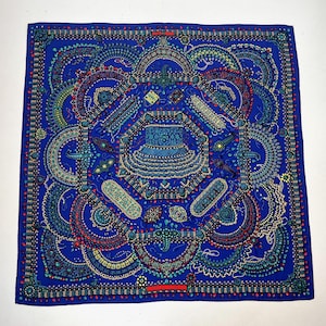 May include: A square, royal blue scarf with an intricate, symmetrical design. The pattern features a central motif surrounded by ornate, dotted lines and shapes in shades of blue, teal, red, and yellow. A red rectangular accent is visible on the bottom edge.