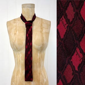 May include: A vintage necktie with a diamond pattern in shades of red and black is displayed on a mannequin. The tie's design features a repeating pattern of red diamonds against a darker background. The tie is knotted at the neck.